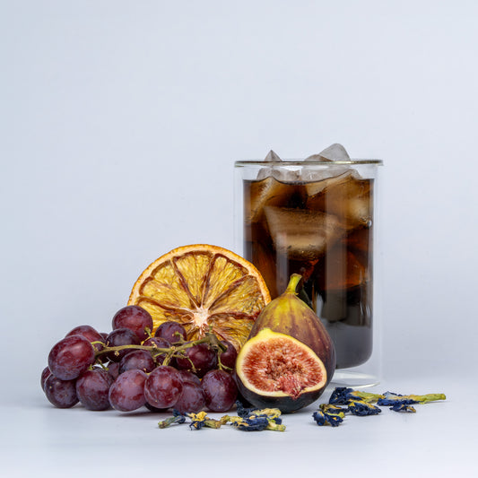 Glass of iced coffee with grapes, figs, and a dried orange on a light gray background